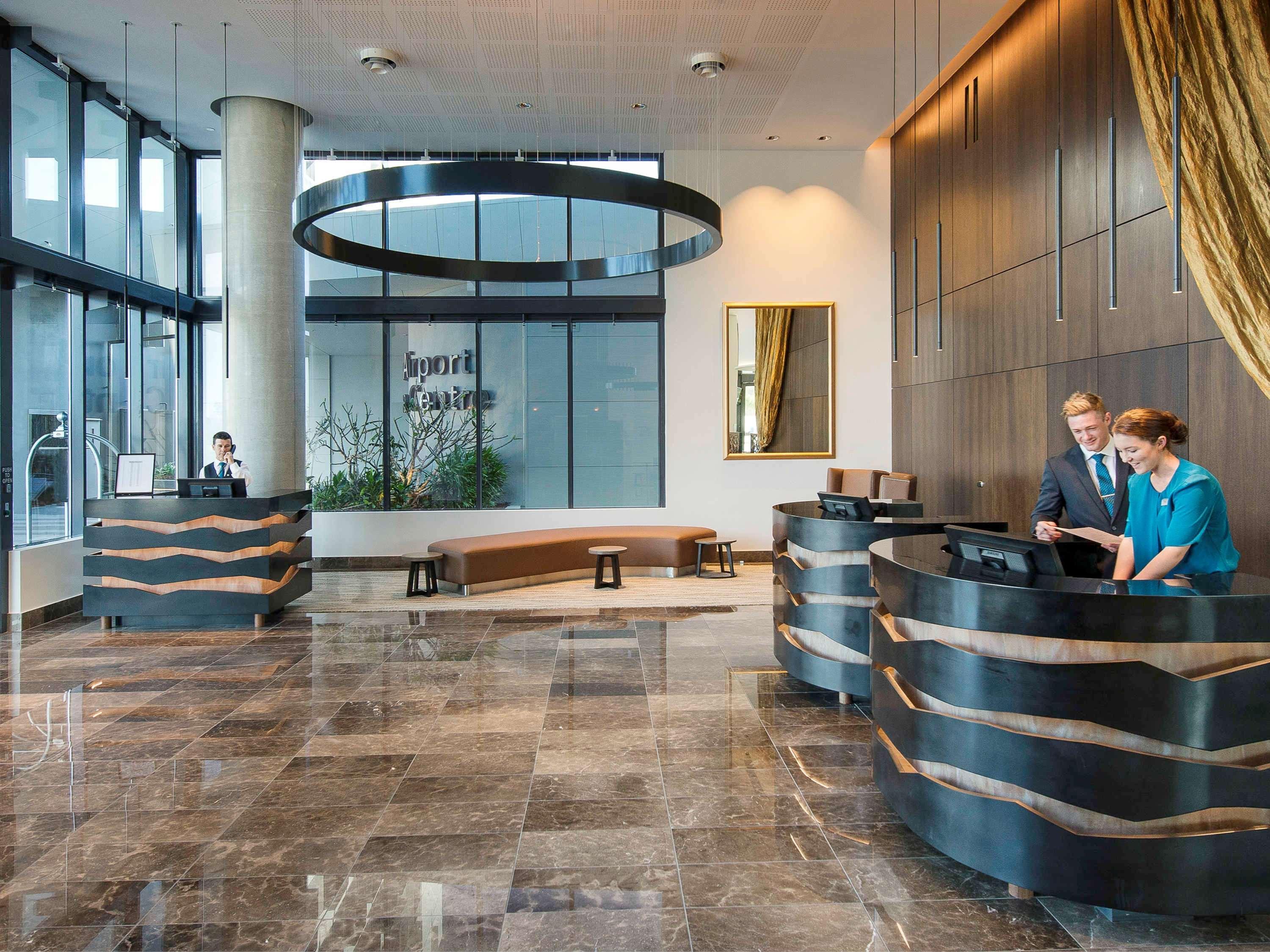 Pullman Brisbane Airport Hotel Bagian luar foto The image shows a modern hotel lobby. There are two reception desks made of dark-colored materials, and staff members are attending to guests at these desks. The lobby features large windows that allow natural light to fill the space, and there are p